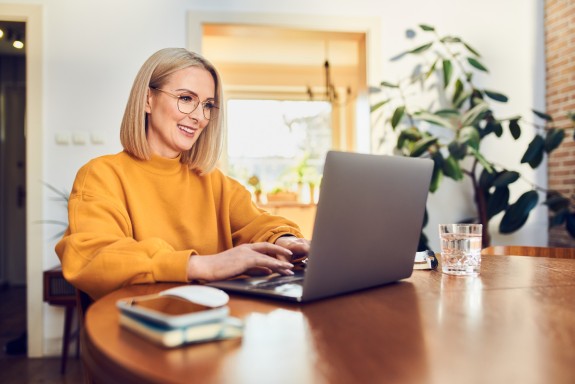 blonde Frau mit Brille und gelbem Pullover sitzt daheim am Laptop